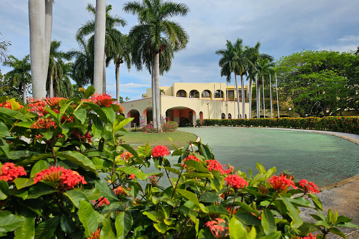 Exterior architecture of the Sevayu Ayurveda Resort in Havana, Cuba, featuring tropical palm trees and vibrant red Ixora flowers in the foreground.